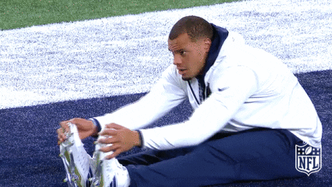 An animated gif of a football player sitting on the field doing a forward fold stretch, he's pulling his feet back one at a time so it almost looks like a dance