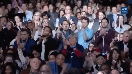 Video gif. Crowd of people in business attire slowly rise from their seats and clap fervently. Jill and Joe Biden and Michelle Obama pop up from the front row.