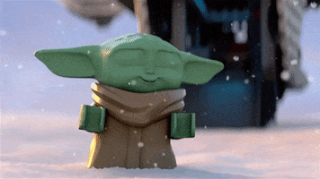 Movie gif. Baby Yoda from Lego star wars hops up and down in the snow. He excitedly looks at the snow that’s falling all around him. 