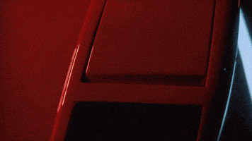 Music video gif. From Octavian's "Rari," a close-up of a red Ferrari's front headlight popping open, panning around to the driver side where a man bobs his head behind the wheel.