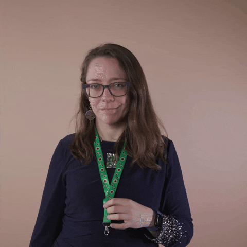 Reaction gif. A Disabled Latina woman with brown wavy hair and glasses shows off her green lanyard, presenting it with an ok sign and a knowing smile.