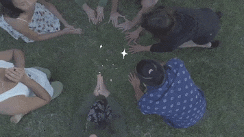 Video gif. Six people kneel in a circle and then sit up to pray. Sparkles glitter in the center of their prayer circle.
