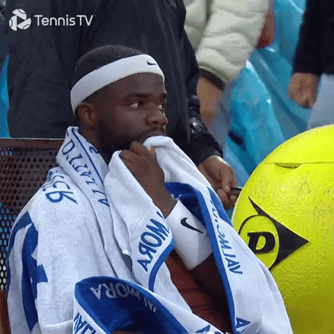 Sports gif. Frances Tiafoe sits on a bench at a game with a towel over his shoulders and a sweatband on his head. He pulls the towel to his mouth, looking off with a concerned expression.