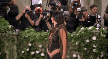 Met Gala 2024 gif. Kendall Jenner wearing a plunging black and bronze vintage Givenchy 1999 Haute Couture gown posing over her shoulder, surrounded by paparazzi.