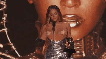 Video gif. Beyonce holds a Grammy award in her hand as she stands at a microphone and tosses her hair over her shoulder, looking up and giving a quick sigh as if she's collecting herself in the moment to give an acceptance speech. 