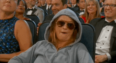 TV gif. Amy Poehler sits in a crowded auditorium wearing a hoodie and sunglasses. She chews gum with a chill, stoic expression while throwing double peace signs, as if telling us "peace, I'm out".