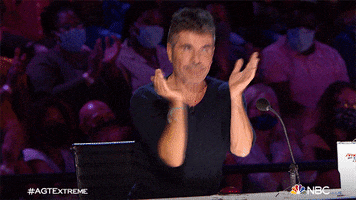 Reality TV gif. Simon Cowell as judge on AGT stands up from his table, claps, and gives two thumbs up to right of frame.