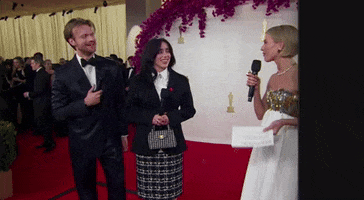 Oscars 2024 GIF. Julianne Hough interviews Finneas and Billie Eilish on the red carpet and Eilish laughs. Eilish and Finneas wear black suits and both wear matching ceasefire pins.