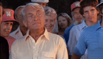 Movie gif. Ted Knight as Judge Smails in Caddyshack stands in front of a crowd of people. His eyes are wide and he shakes his head sarcastically as he says, “Well? We’re waiting!”