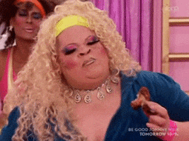 Reality TV gif. A drag queen from RuPaul's Drag Race is eating a chicken wing and they're living, shaking their chest and shoulder in excitement.
