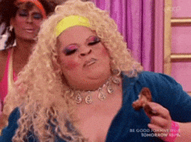 Reality TV gif. A drag queen from RuPaul's Drag Race is eating a chicken wing and they're living, shaking their chest and shoulder in excitement.