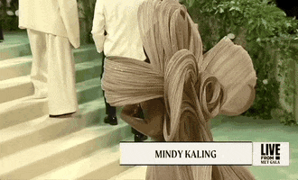 Met Gala 2024 gif. Mindy Kaling turns to reveal the back of her champagne-colored Gaurav Gupta gown, showing the dramatic floaty sheer loops of sculptural fabric that form a giant loose knot at her upper back. The loose wavy pattern flows through a long train that trails behind her.