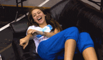 Celebrity gif. Miley Cyrus points at someone and kicks up her legs as she laughs on a couch.