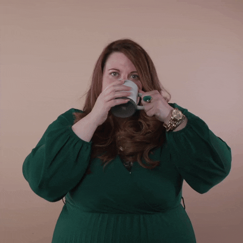 Reaction gif. A non-apparently Disabled white woman with with anxiety and depression and long red hair taking a sip from her mug, takes a moment to smile a wave at us enthusiastically, saying, "Good morning!"