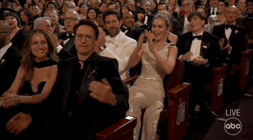 Oscars 2024 GIF. Robert Downey Junior and Susan Downey, Emily Blunt and John Krasinski, Cillian Murphy, and Ben Kingsley seated in the aisle seat at the Oscars, applaud, RDJ pumping his fist at his heart to punctuate self-confidence, then dropping the act and shrugging it off.