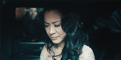 Movie gif. Michelle Yeoh as Movie Star Universe Evelyn in Everything Everywhere All at Once, dressed glamorously for a red carpet event, squints, her eyes adjusting, as she prepares to exit her limousine, flash bulbs going off around her.