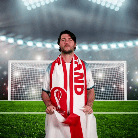 Video gif. Man stands in front of a soccer field with a goal and stadium lights behind him. He holds a British flag up and screams like a serious fan, "Let's go, Englandaaand!'