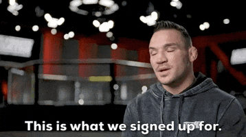 Video gif. Michael Chandler on The Ultimate Fighter sitting for in an interview shrugs and says, "This is what we signed up for."