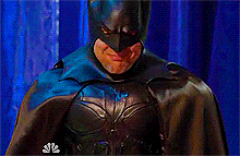 Video gif. Man in a Batman costume scrunches his face in a frown as he bows his head and squeezes the bridge of his nose.