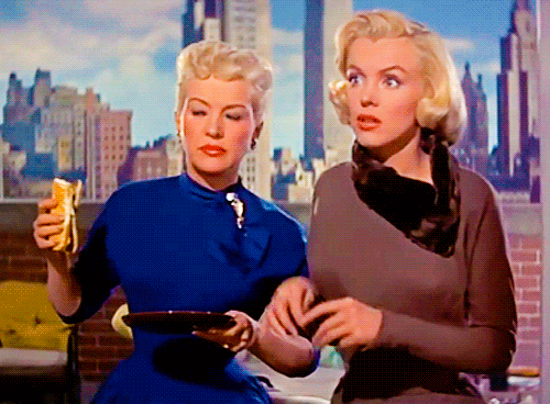 Movie gif. Marilyn Monroe as Pola in "How to Marry a Millionaire" raises her glasses to her face for a better look while Betty Grable as Loco looks on while eating a sandwich.