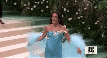 Met Gala 2024 gif. Lea Michele wearing Rodarte, an aqua gown with 3D white flowers across one shoulder an oversized tulle cape worn off her shoulders, empire-waist showing off her baby bump, in slow motion she throws her arms out in a welcoming gesture that says "I've arrived," a big smile on her face.
