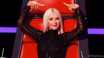 Reality TV gif. Christina Aguilera on the Voice sits in her chair and uses both hands to point to herself, wiggling her fingers like worms to emphasize that she wants to be picked. She has a smug smirk on her face as if she knows she’s the right choice.