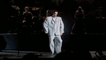 Celebrity gif. David Bryne is dancing on stage in a giant, oversized suit with wide shoulder pads. He wiggles his legs and flaps his arms, emphasizing the largeness of his suit.