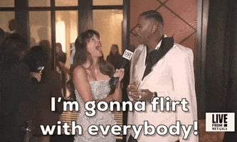 Met Gala 2024 gif. Colman Domingo laughs with a reporter, saying "I'm going to flirt with everybody." The reporter cracks up in response. Doming wears an oversized, white-and-black Willy Chavarria suit with diamond earrings and a bouquet of white flowers wrapped in a white satin cloth. This is paired with a full-length ivory colored cape that hangs from the shoulders and sweeps back in a short train. 