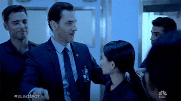 TV gif. Man wearing a badge gives his colleagues a smile and two thumbs up as if to say, “Good job!”