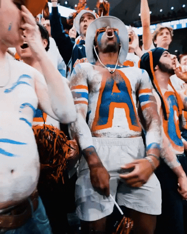 Sports gif. Auburn Tigers basketball fan has an A painted in the school colors onto his bare chest and is roaring in support. Another fan appears in an all white suit jumping up and down throwing his into the air. They are surrounded by an excited crowd of students at an Auburn game. 