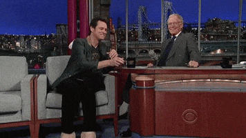 Late Show gif. In a scene with David Letterman, Jim Carrey sits in an armchair next to David's desk. He takes a coffee mug from the desk and leans back, then kicks up a pair of fake oversized bare feet onto the corner of the desk as he casually sips from the mug.