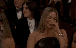 Oscars 2024 GIF. Margot Robbie, seated at the Oscars, covering her mouth, snickers and giggles uncontrollably.