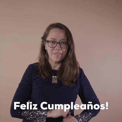 Reaction gif. A Disabled Latina woman with brown wavy hair and glasses smilingly explodes her jazz hands in an outward arc, shouting "Feliz cumpleanos!"