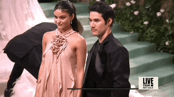 Met Gala 2024 gif. Camila Mendes poses with Joseph Altuzarra. Mendes is wearing a dusty rose halter dress with a rosebud detailing where the straps meet at the center of her chest. The dress has delicate flowing vertical panels that hang straight. Altuzarra is wearing a black shiny suit jacket and shirt.