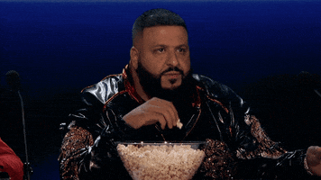Celebrity gif. DJ Khaled sits with a bowl of popcorn in front of him. He stares at something almost like he’s entranced as he shovels handfuls of popcorn into his mouth.