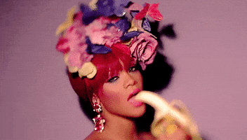 Music video gif. In the video for S&M, Rihanna wears a flower crown and makes eye contact us while slowly bringing a banana to her mouth.