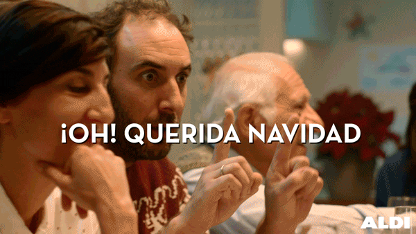 Video gif. A man with brown hair and a beard, sits near a woman. He raises one finger on each hand, then hits his hands together, revealing two fingers raised on one hand and closed fist on the other. He raises his eyebrows at his magic trick. Text, in Spanish "¡Oh! Querida Navidad."