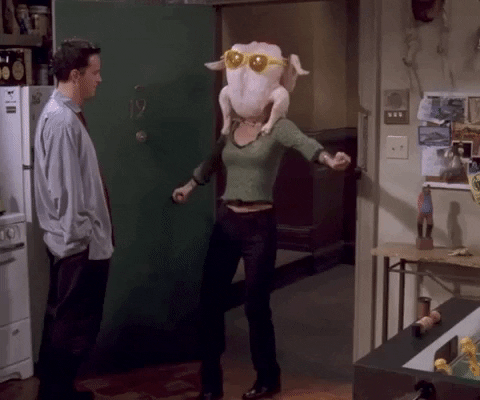Friends gif. Courteney Cox as Monica wears a giant turkey with sunglasses on her head as she shimmies through an apartment door. Matthew Perry as Chandler looks on casually with his hands in his pockets. 
