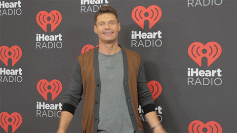 Celebrity gif. Ryan Seacrest waves both hands in front of an iHeartRadio backdrop and says, “Hi!”