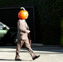 Video gif. A man wearing a suit and carrying a briefcase walking through a parking lot with a Jack-O-Lantern on his head.