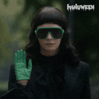 Sponsored gif. Woman wearing futuristic green sunglasses and matching leather gloves waves one hand and just barely smiles at us.