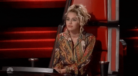 Reality TV gif. Miley Cyrus as a judge on The Voice. She leans back in her seat and looks eager as she smiles and says, "Yes!" while clapping her hands.