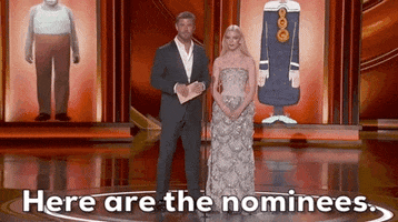 Oscars 2024 gif. Chris Hemsworth and Anna Taylor Joy stand at the microphone onstage wearing a gray tuxedo and a nude-colored dress adorned with silver sparkles. Taylor Joy bends ever so slightly and says, "Here are the nominees."