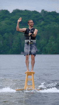 Video gif. A smiling man wakeboarding on a barstool attached to a tabletop waves his hand in a cupping motion, like the queen.