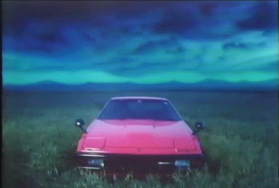 Video gif. Flashy 1980s red sports car sits alone in a vast grassy plain underneath a mysterious sky with a blanket of dark rolling clouds and a bright turquoise glow.
