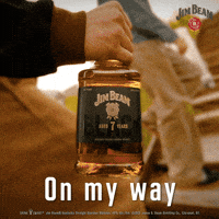 Sponsored gif. Close up of a bottle of Jim Beam Black bourbon whiskey as it’s carried up a flight of outdoor stairs by someone. Text reads, “On my way.”