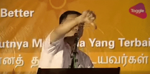 Video gif. Man stands at a podium and he angrily shoves a big thumbs down towards the crowd as he says, “Boo” into the microphone.