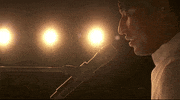 Oscars 2024 GIF. Close up shot of Jon Batiste performing "Never Went Away" from American Symphony. He's shot from the stage angle and we see the lights of the audience shining brightly behind him as he sits in a glowing halo of yellow light. 