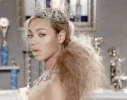 Music video gif. Beyonce wears a sparkly crown hanging off her head and a big poofy messy teased ponytail. She shimmies her shoulders as she looks at us, chewing on gum against a background filled with children's sports trophies