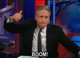 TV gif. Jon Stewart on the Daily Show spins in his chair at his onstage desk, yelling, "Boom!"
